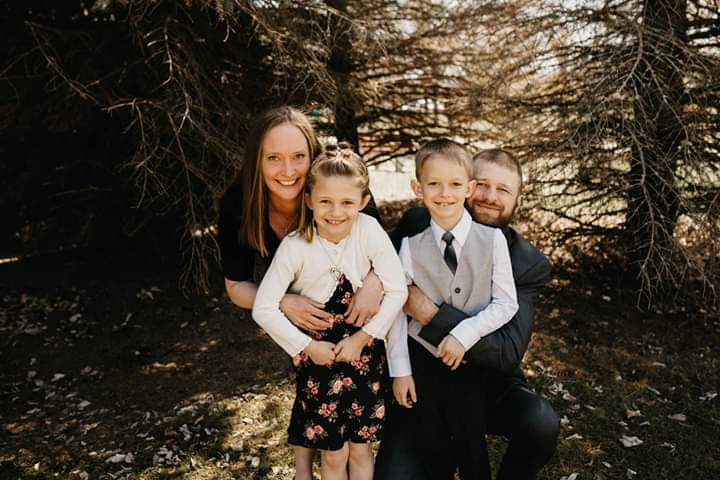 Family portrait featuring a woman, two children, and a man outdoors. The woman is wearing a black shirt, the girl has a floral dress, and the boy is dressed in a suit. They are smiling and posing together in front of a backdrop of trees.
